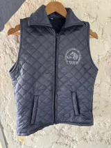 Chaleco acolchado azul marino con diseño pespunteado en rombos, cuello alto y cierre frontal con cremallera. Presenta un bordado institucional en el pecho y bolsillos laterales.