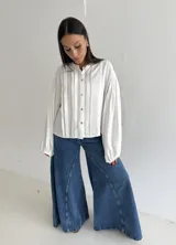 Blusa blanca con cuello mao y cierre frontal de botones de nácar. Presenta mangas largas abullonadas con puños elásticos y detalle de tablas verticales en el frente.