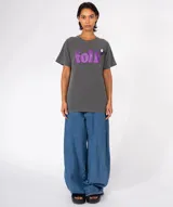 Remera de algodón color gris oscuro con lavado especial, corte holgado y cuello redondo. Estampa frontal con la palabra "folk" en glitter morado. Lleva un pin decorativo en la zona del hombro.