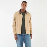 Chaqueta de trabajo (chore coat) color beige, confeccionada en tejido de algodón tipo sarga o drill, con cuello contrastante de pana marrón oscuro. Presenta cierre frontal de botones metálicos y cuatro bolsillos frontales de parche (dos superiores y dos inferiores).