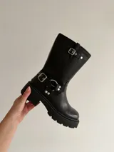 Bota de caña media estilo biker, confeccionada en eco cuero negro. Presenta suela track con plataforma, puntera redondeada y detalles de correas con hebillas metálicas y argollas decorativas en el tobillo.