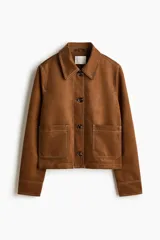 Chaqueta corta marrón de acabado cepillado suave con costuras en contraste. Cuenta con cuello clásico, cierre frontal de botones, bolsillos de parche delanteros y mangas largas.