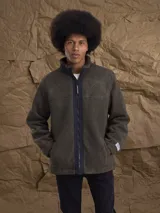 Campera de sherpa color verde oliva con cierre frontal completo, cuello alto y detalles en contraste negro en la cremallera. Presenta una etiqueta decorativa en el puño.