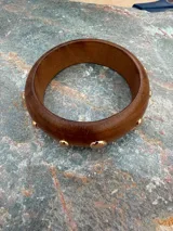 Pulsera rígida de madera marrón con tachas metálicas doradas distribuidas a lo largo de su contorno.