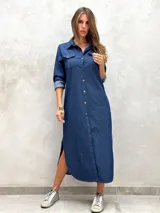Vestido camisero largo de denim azul oscuro, con cuello camisero, botones frontales y bolsillos de parche en el pecho. Presenta mangas largas remangadas y aberturas laterales en el ruedo.