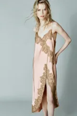 Vestido largo tipo lencero de satén color rosado, con escote en pico y tirantes finos. Presenta aplicaciones de encaje a contraste en el escote y en el lateral, con un diseño de bajo asimétrico.
