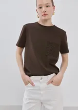 Remera marrón de corte recto, con cuello redondo, manga corta y detalle de bolsillo con bordado en el pecho.