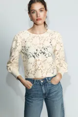 Camisa de encaje color crema con cuello redondo, mangas largas abullonadas y puños ajustados. Presenta cierre en la espalda con botones dorados.