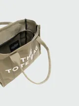 Bolso tipo tote de Marc Jacobs, confeccionado en lona resistente de color negro. Presenta el nombre del modelo estampado en blanco en la parte frontal y el logo de la marca debajo. Cuenta con doble asa superior para llevar al hombro o en la mano.