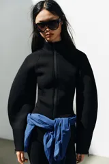 Chaqueta negra de cuello subido con cierre frontal de cremallera termosellada, manga larga con puños anchos y cremalleras laterales en los puños.