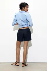 Bermuda de tiro medio color azul marino, con pinzas delanteras, cierre frontal y cinturón fino color beige.