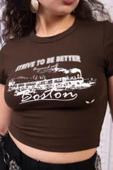 Remera marrón de corte ajustado tipo baby tee, manga corta y cuello redondo, con estampado frontal en blanco de un tren y la leyenda 'Strive to be better Boston'.