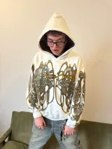 Hoodie oversize de color blanco con estampado artístico en tonos marrones y dorados en el pecho y las mangas. Presenta un forro interior de capucha en tejido marrón texturizado y costuras reforzadas.