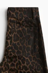 Pantalón de jean de tiro alto con pierna amplia y corte holgado, confeccionado en tela con estampado de leopardo en tonos marrón y negro. Presenta cierre y dos botones dorados en la cintura, y pliegues frontales.
