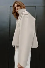 Trench corta de color beige, con cuello solapa, mangas largas con trabillas en los hombros, bolsillos delanteros de vivo y cierre frontal cruzado con botones a presión.