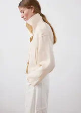 Chaqueta crop de lino color beige, diseño recto, cuello alto tipo embudo, manga larga, dos bolsillos laterales y cierre cruzado de doble botonadura metálica dorada con detalles en los hombros.