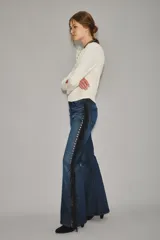 Jean de corte recto y tiro medio, color azul oscuro, con detalle de flecos negros y ojales metálicos a lo largo de la costura lateral.