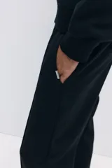 Pantalón jogger negro de corte regular fit, confeccionado en tejido suave con mezcla de algodón. Presenta pliegues cosidos en la parte delantera, cintura elástica con cordón ajustable, bolsillos laterales y un bolsillo insertado en la parte trasera.