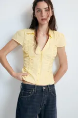 Top de cuello solapa y escote en pico, con manga corta y cierre frontal con botones. Presenta un diseño ajustado con detalles de frunces en el delantero.