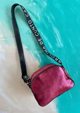 Bandolera Kaira de la línea Ottenta de Carolina De Cunto, en color fucsia metalizado. Cuenta con un diseño compacto, bolsillo exterior con broche imantado y detalle pintado a mano, cierre superior y una correa larga regulable con el logo de la marca estampado.