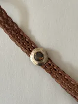 Cinto trenzado de cuero color camel con hebilla circular metálica grabada y detalle de piedra central.