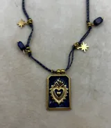 Collar con cordón trenzado azul oscuro con hilos dorados, dije rectangular dorado con un sagrado corazón en relieve sobre fondo azul esmaltado. Incluye cuentas de lapislázuli y dijes colgantes dorados con forma de estrella.