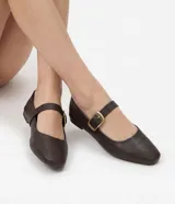 Zapatilla tipo Mary Jane de cuero marrón, con punta redondeada y tira en el empeine con hebilla metálica dorada.