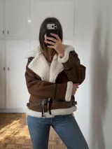 Campera tipo gamulán de color marrón con interior y solapas de corderito sintético en color crema. Presenta cierre frontal, detalles de tiras en cuero sintético negro en puños, cuello y ruedo, y hebillas metálicas ajustables.