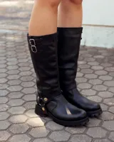 Bota de caña alta confeccionada en cuero negro, con suela track de goma. Presenta un diseño versátil con dos hebillas ajustables en la parte superior de la caña y una correa decorativa con tachas metálicas y argollas en la zona del tobillo.