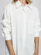 Camisa blanca de corte oversize, con cuello clásico, cierre frontal con cartera oculta y bolsillo delantero. Presenta mangas largas con puños abotonados.