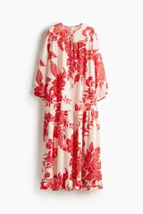 Vestido largo de tejido ligero color blanco con estampado floral rojo. Tiene cuello redondo con lazos, mangas globo largas con puños abotonados, corte holgado y falda con volantes en el ruedo.