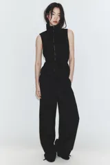 Chaleco negro de diseño fluido con cuello subido y cierre frontal de cremallera metálica. Presenta cordones ajustables con lazadas en el cuello y en la cintura.