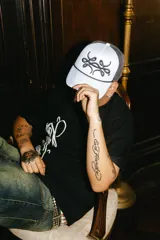 Gorra trucker gris con paneles frontales blancos y paneles traseros de rejilla negra. Presenta un logo bordado en el panel frontal y una cuerda decorativa en la base de la corona.