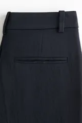 Pantalón sastrero de tiro alto con cierre, botón y corchete ocultos. Presenta piernas rectas con pliegues frontales y traseros, bolsillos laterales al bies y bolsillos traseros ribeteados.