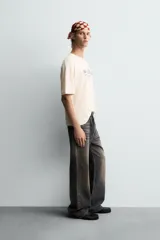 Camiseta de corte boxy fit, color beige con efecto lavado, cuello redondo y manga corta. Presenta un estampado gráfico en azul oscuro en el frente con tipografía estilo vintage.