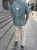 Remera verde con estampa en la espalda que dice "Malibu Sports Club" con una palmera en el centro y la frase "Livin' The Lifestyle" abajo.