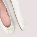 Bailarinas de charol blanco con lazo ajustable en la puntera, suela de goma antideslizante, plantilla de piel con memory foam y forro de tela.