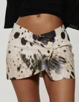 Minifalda de cuerina con estampado animal print tipo vaca en tonos crudo, negro y marrón, con diseño cruzado asimétrico y botones metálicos decorativos en el frente.