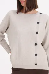 Sweater de tejido de punto color beige, con cuello redondo y detalle de botones negros decorativos en el frente. Presenta mangas largas con puños acanalados y un corte recto.