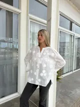 Camisa blanca de corte holgado con cuello mao y cierre frontal de botones. Presenta un diseño bordado con motivos florales en toda la prenda y mangas largas con puños amplios.