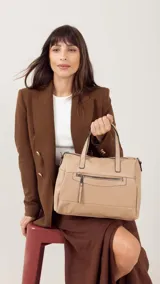 Cartera de mano y hombro color beige, de material símil cuero, con forma estructurada y redondeada. Posee dos asas cortas superiores y una correa larga ajustable y desmontable. Cuenta con un bolsillo exterior con cierre en la parte frontal.