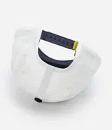 Gorra blanca de sarga con visera azul marino y bordado de Snoopy jugando baseball.
