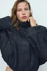 Cazadora de denim color azul oscuro, con cuello solapa, mangas largas tipo murciélago y bolsillos de vivo en el delantero. Presenta un bajo elástico ajustable con cordones y cierre frontal con cremallera y botones a presión ocultos.