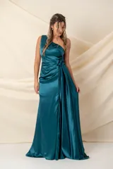 Vestido largo de satén color verde petróleo, con diseño asimétrico de un solo hombro y abertura frontal en el escote. Presenta una cintura drapeada con detalle de flor lateral, falda con tajo frontal y cierre de botones forrados en la espalda.
