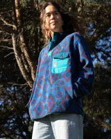 Buzo de polar fleece con diseño estampado geométrico en tonos azules, bordó y verde. Presenta cuello alto con cierre de botones a presión, bolsillo frontal con solapa en contraste color turquesa y puños elásticos.