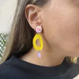 Aros colgantes con pasador de acero quirúrgico, compuesto por un círculo superior blanco con detalles en rosa y amarillo, un dije calado amarillo en forma de gota y un dije inferior en forma de lágrima color lila.