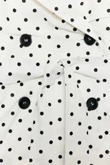 Cazadora estilo trench corta confeccionada en lino, con diseño de lunares negros sobre fondo blanco. Cuenta con cuello solapa, manga larga con trabillas en los puños, cierre frontal cruzado con botones negros y cinturón ajustable del mismo tejido.