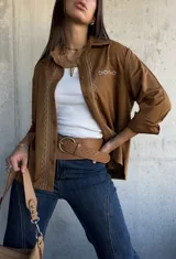 Camisa de gamuza color camel, con cuello clásico, cierre frontal de botones y detalle de bordado geométrico en color crema a lo largo de la botonadura y en el bolsillo frontal. Presenta mangas largas con puños doblados.