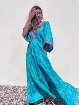 Túnica larga de estilo boho, color turquesa con estampado paisley en tonos azules y dorados. Presenta escote en V profundo con bordados decorativos en color fucsia y blanco, mangas amplias tipo kimono y ajuste en la cintura con cordón. El diseño incluye un ruedo con volado y terminaciones con borlas.