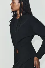 Chaqueta de cuello redondo y manga larga con pliegues, de color negro, con falsos bolsillos de vivo en el delantero y cierre frontal con botones.
